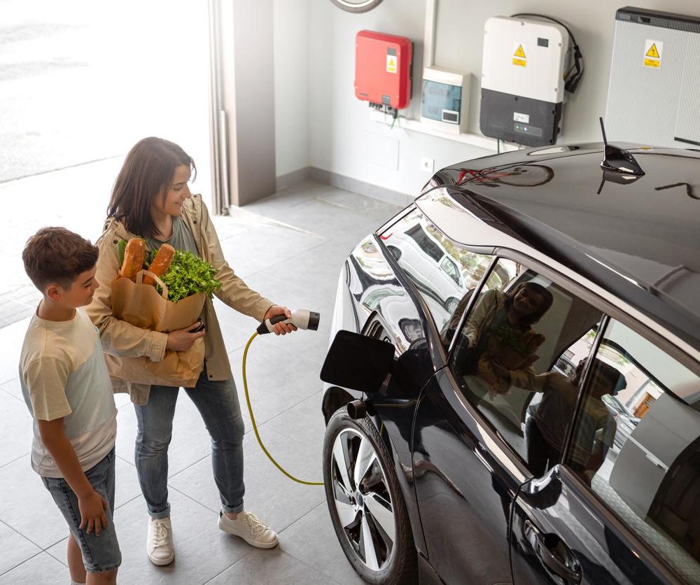 Happy son and mother plug EV charger from home charging station to electric vehicle. Future eco-friendly car powered by renewable source of clean energy on daytime. Horizontal, high angle.